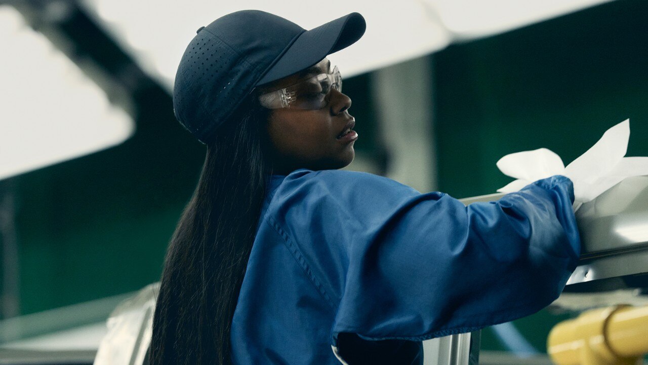 Automotive worker wearing protective gear and inspecting a vehicle component in a factory environment.