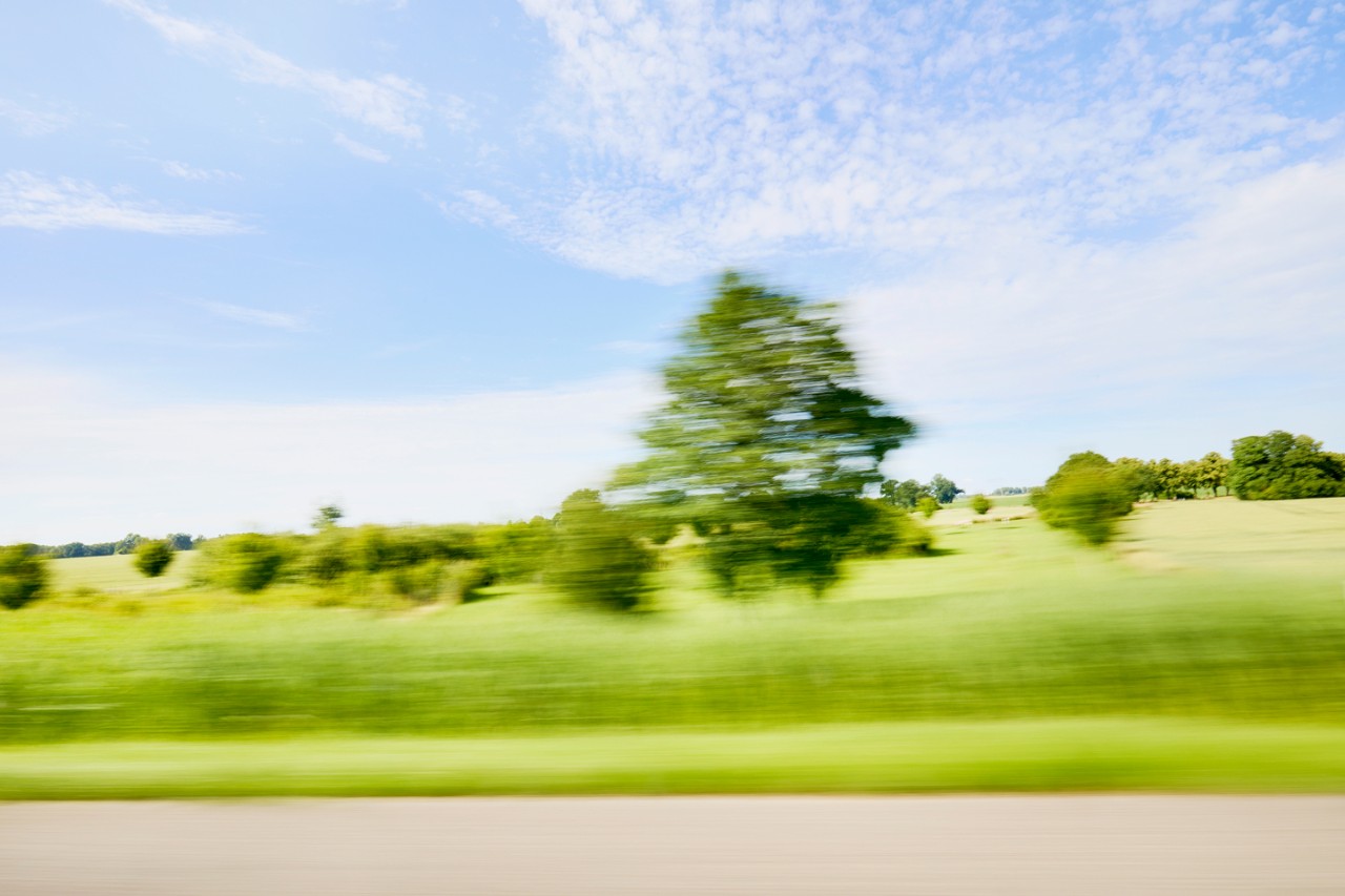 A blurred view of greenery and a blue sky from a car in motion on the road.