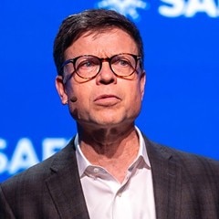Kurt Kelty wearing a black suit and presenting on stage at a SAFE event, standing in front of a branded blue background.