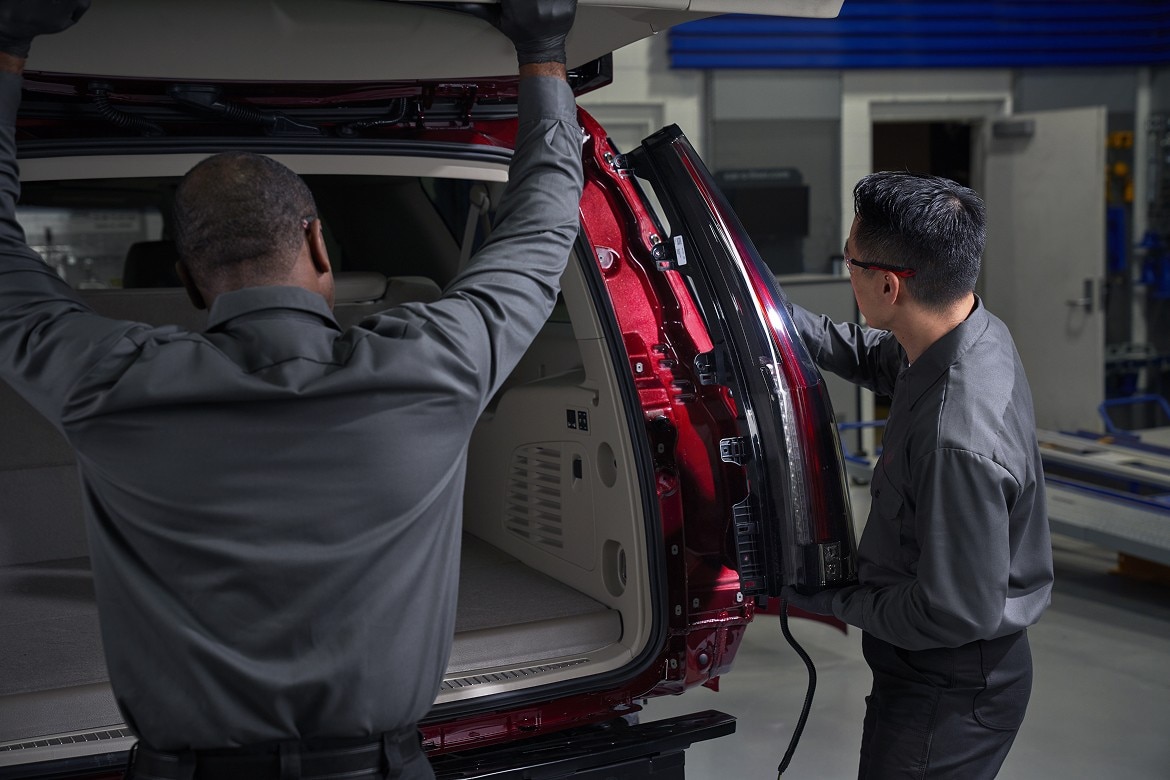 Two automotive technicians inspect the assembly of a red SUV with its rear liftgate open, inside a manufacturing facility.