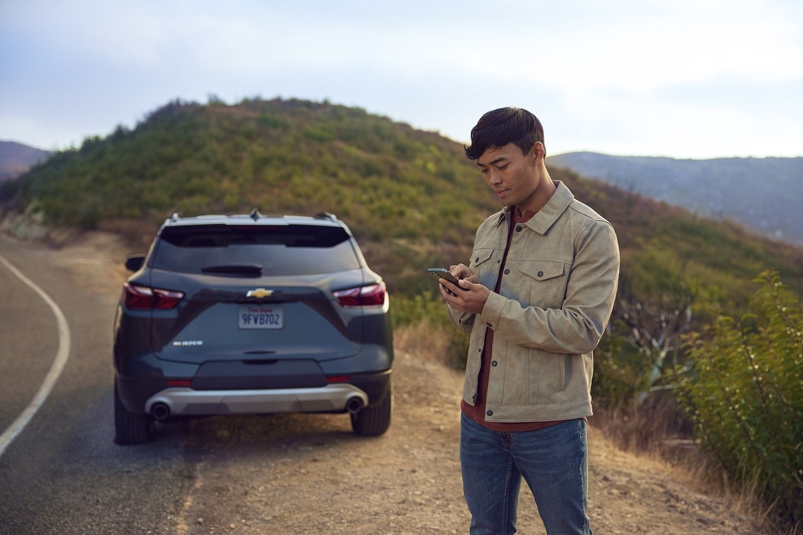 Man uses his smartphone standing beside a parked Chevrolet SUV on a scenic roadside with mountains and greenery.
