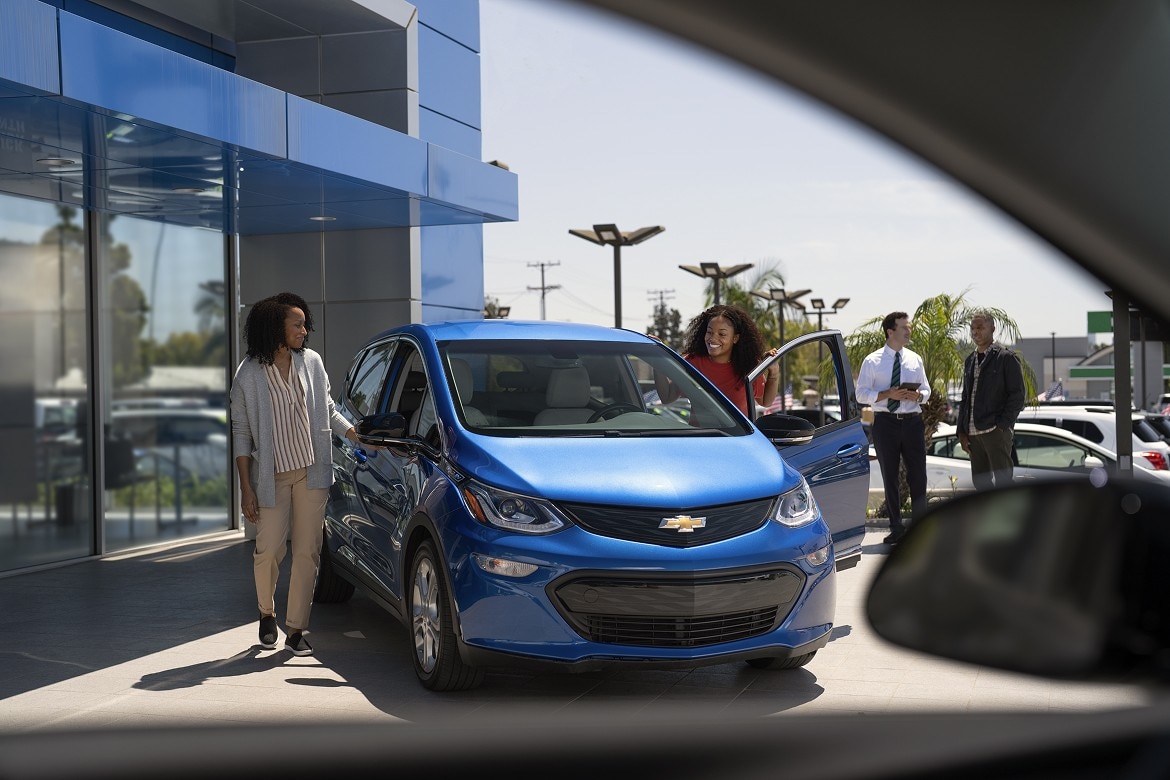 Two women examine a blue Chevrolet EV outside a dealership. Two salesmen talk beside palm trees in the background.