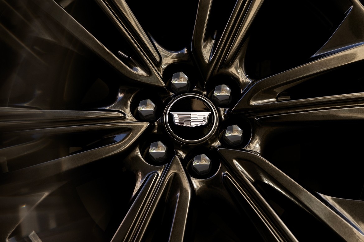 Close-up of a glossy black alloy wheel with the Cadillac emblem centered among polished lug nuts, reflecting the surrounding.